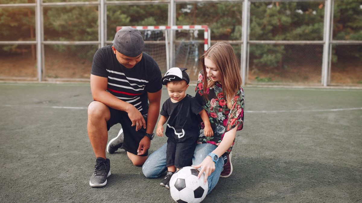 Full body of positive smiling family with cute little boy resting on football field with ball against football gate